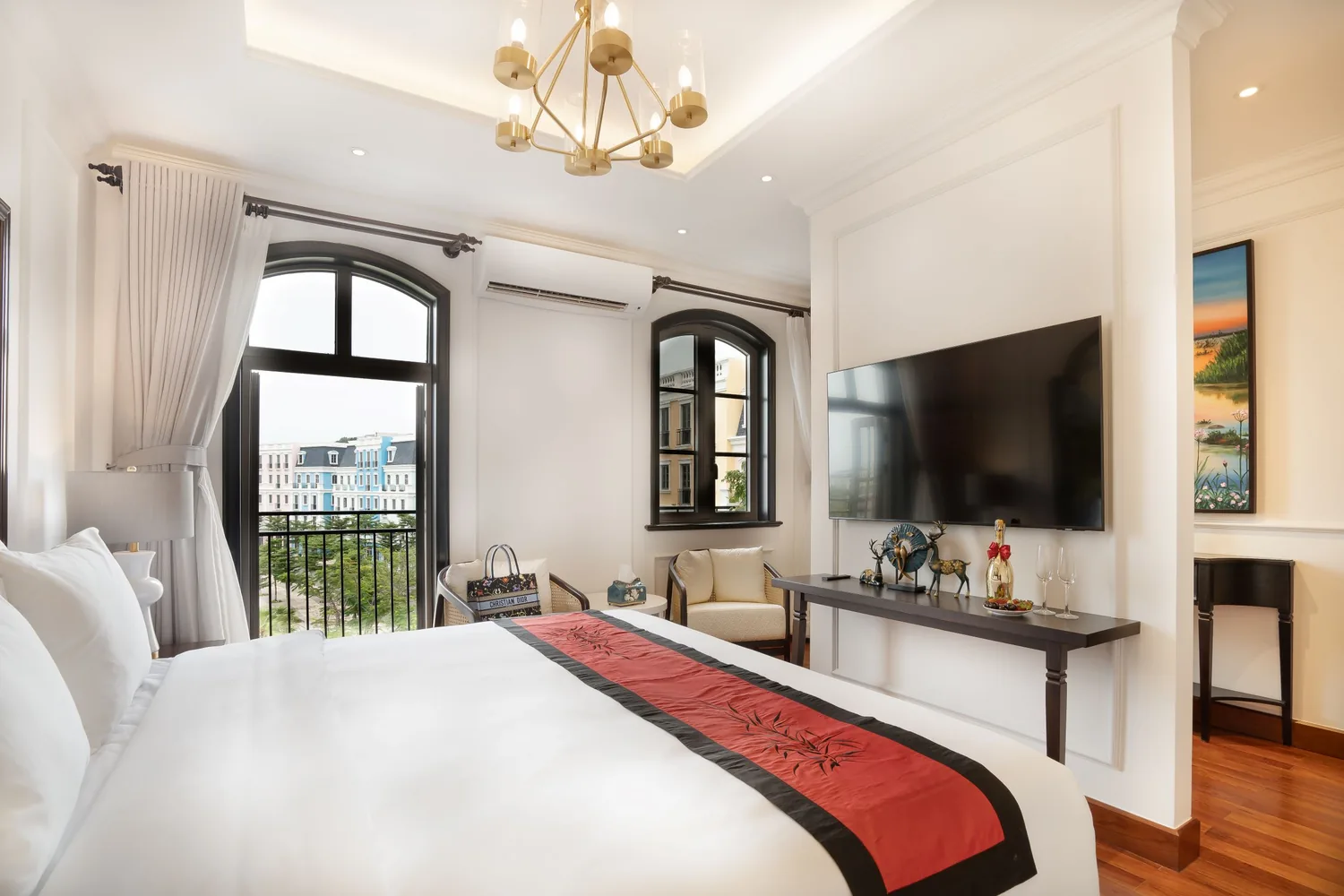 La Rose Suite bedroom with arched French windows opening to a balcony over Sunset Town, a brass chandelier and a red bamboo-motif runner